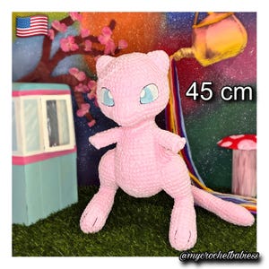 May include: A light pink crocheted plush toy featuring large blue eyes and a long tail. The figure stands upright and is labeled with a height of 45 cm. The plush has a soft, textured yarn surface and is positioned in a standing pose with arms extended.