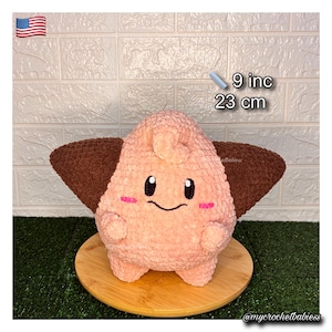 May include: A plush, light pink crochet doll with brown ear-like wings. The doll has a smiling face with black eyes and pink cheeks. It measures 9 inches in height. The doll is sitting on a wooden surface.