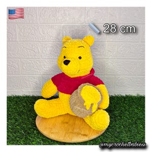Puede incluir: Un peluche de oso tejido a ganchillo en amarillo y rojo que sostiene un tarro de miel color canela con miel amarilla goteando desde la parte superior. El peluche mide 28 cm de altura y tiene cabeza redonda, orejas pequeñas y nariz negra.