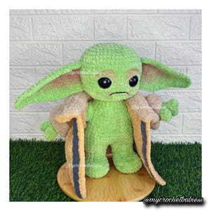 May include: A green crocheted plush toy of a character with large ears and black eyes, wearing a tan and black coat. The toy is standing on a wooden base. The text "@mycrochetbabiess" is visible.