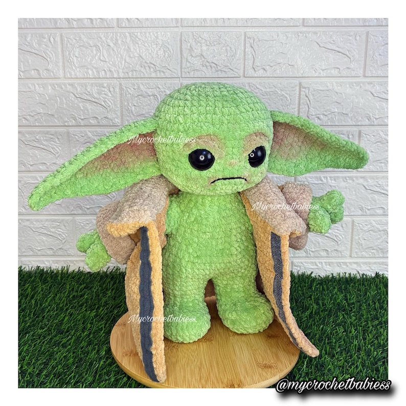 May include: A green crocheted plush toy of a character with large ears and black eyes, wearing a tan and black coat. The toy is standing on a wooden base. The text "@mycrochetbabiess" is visible.