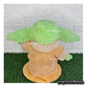 May include: A crocheted figure of a character with large green ears and a tan robe. The figure is sitting on a wooden base. The figure is made of soft, textured yarn. The figure is a popular character from a science fiction franchise.