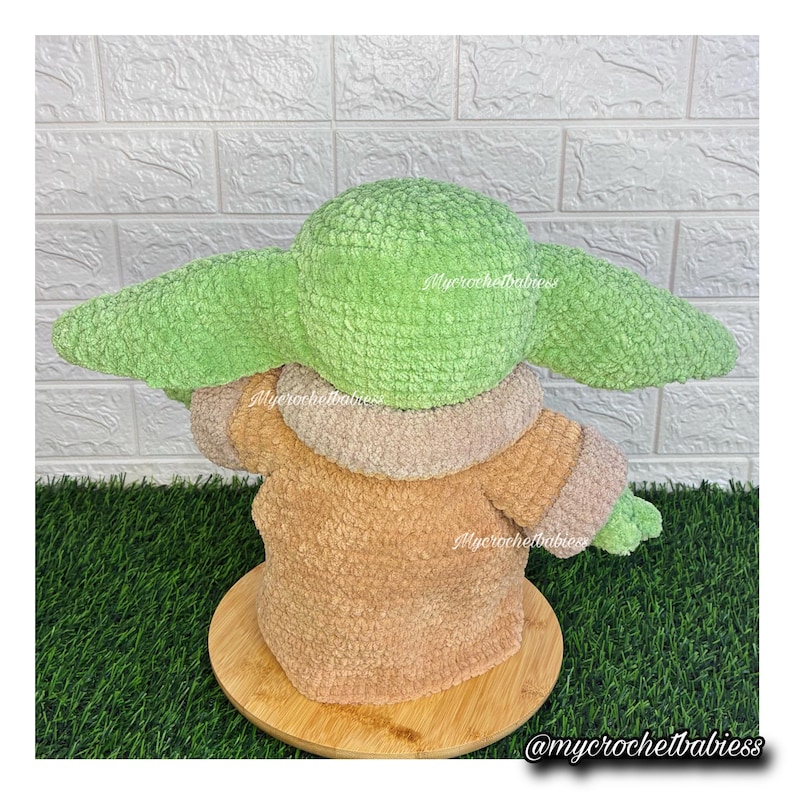 May include: A crocheted figure of a character with large green ears and a tan robe. The figure is sitting on a wooden base. The figure is made of soft, textured yarn. The figure is a popular character from a science fiction franchise.