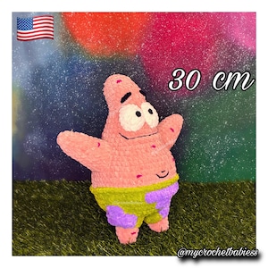 Pode incluir: Um peluche de croché cor-de-rosa em forma de estrela-do-mar com olhos brancos grandes, sobrancelhas pretas e uma boca sorridente. A figura usa calções com padrão verde-lima e roxo e mede 30 cm de altura. A textura é macia e de fio.