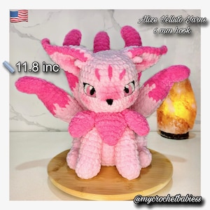 May include: A handmade pink and white crochet creature, approximately 11.8 inches tall, with large ears and wings. The creature is sitting on a wooden round base. The image includes text that says "Alize Velluto Yarns 6 mm hook" and "@mycrochetbabiess".