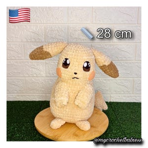 May include: A handmade, plush Pikachu doll, approximately 28 cm tall, in a light yellow color with brown accents on the ears and tail. The doll has black eyes, pink cheeks, and is displayed on a wooden base. The text "28 cm" is visible.