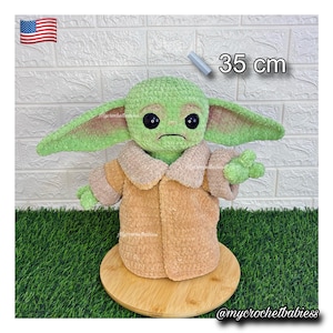May include: A green and tan crocheted doll, 35 cm tall, with large ears and black eyes. The doll is wearing a tan coat and is displayed on a wooden base. The doll is a character from a popular science fiction franchise.