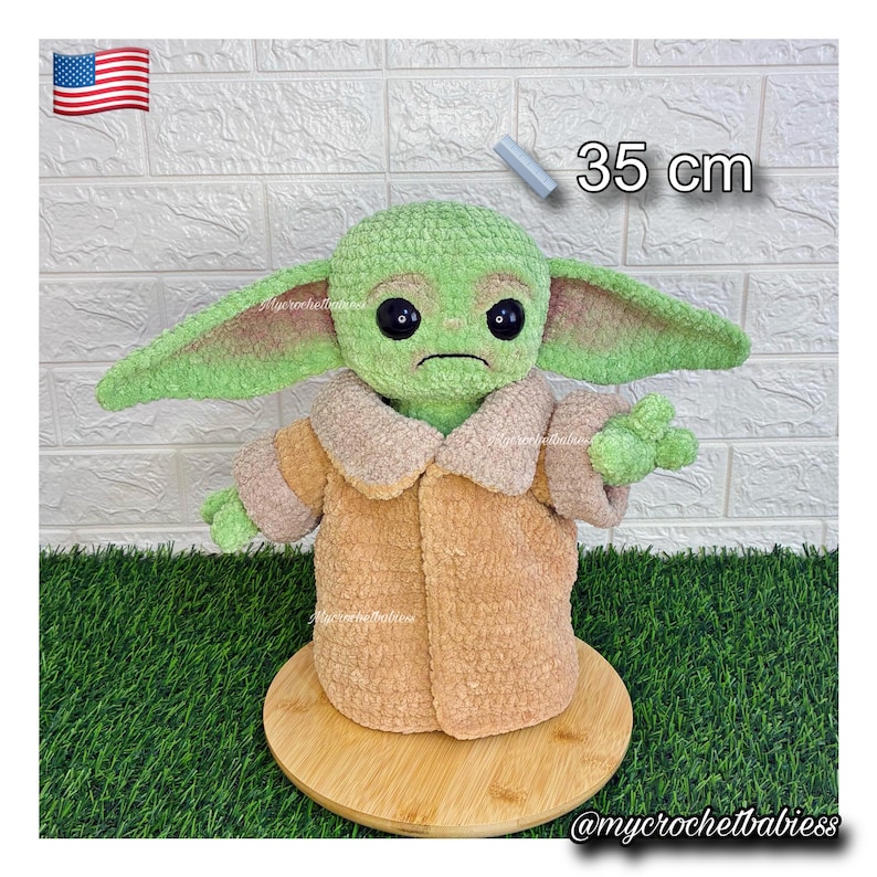 May include: A green and tan crocheted doll, 35 cm tall, with large ears and black eyes. The doll is wearing a tan coat and is displayed on a wooden base. The doll is a character from a popular science fiction franchise.
