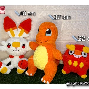 Op de afbeelding: Drie gehaakte pluche speelgoed: een wit en rood konijn (40 cm), een oranje Charmander (37 cm) en een rood en geel wezen (22 cm). Het speelgoed is handgemaakt en staat op een groene ondergrond. Een Amerikaanse vlag staat op de achtergrond.
