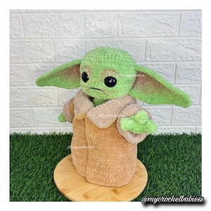 May include: A handmade green and tan crocheted doll, resembling a character from a popular science fiction series. The doll has large ears, black eyes, and is wearing a tan coat. The doll is sitting on a wooden base.