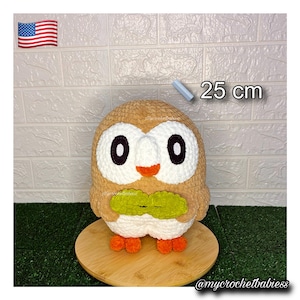 May include: A handmade, plush owl pillow in shades of brown, white, orange, and green. The owl is approximately 9.8 inches tall and has large black eyes and a green leaf-shaped detail. The pillow is displayed on a wooden surface.