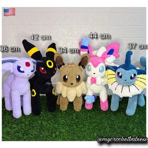 May include: Five plush crochet creatures standing in a row. From left to right: a light purple creature at 36 cm, a black creature with yellow rings at 42 cm, a brown creature at 34 cm, a pink and white creature at 44 cm, and a blue creature at 37 cm.