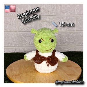 May include: A handmade crochet Shrek doll, approximately 6 inches tall, with a green head, white body, and brown vest. The doll has black eyes and small ears. Text on the image reads "Beginner friendly" and "15 cm".