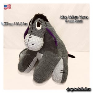 Pode incluir: Um brinquedo de pelúcia Eeyore de crochê feito à mão, com aproximadamente 55 cm de altura. A pelúcia apresenta fios cinza, branco e roxo, com detalhes em preto. O texto "Alize Velluto Yarns 6 mm hook" é visível.