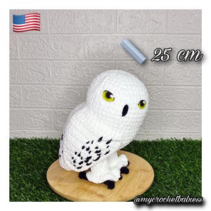 May include: A white crocheted owl plush toy with black wing spots, yellow eyes, and a black beak. The owl is positioned next to a small blue ruler and the text 25 cm, indicating a height of 25 cm. The owl has black feet and a textured yarn finish.