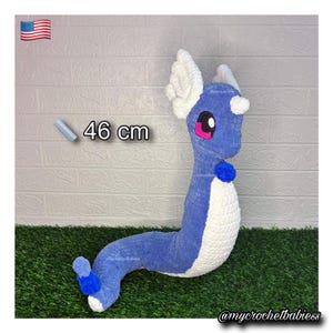 May include: A blue and white crochet plush toy, resembling a dragon-like creature, is displayed against a white brick wall and green grass. The toy is approximately 18 inches long, with pink eyes and a blue pom-pom. The text "46 cm" is visible.
