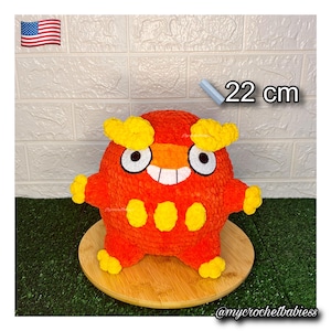 Könnte beinhalten: Ein handgefertigtes, plüschiges, orangefarbenes Wesen mit gelben Akzenten, misst 22 cm. Das Spielzeug hat große, weiß umrandete Augen, einen lächelnden Mund und drei gelbe Punkte auf seinem Körper. Das Spielzeug wird auf einer Holzoberfläche präsentiert.