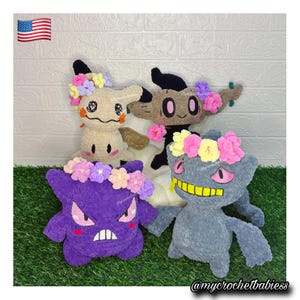 May include: Four plush toys featuring crocheted characters with soft textures. One beige character, one brown tree-like figure, one purple character with a crown of flowers, and one grey character with a yellow mouth and a flower crown.