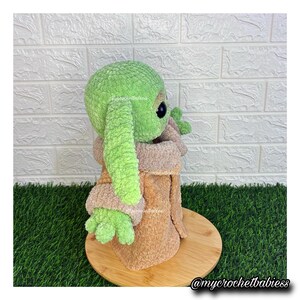 May include: A handmade green and tan plush doll of a character with large ears and big eyes. The doll is wearing a tan robe and is sitting on a wooden base. The doll is made of soft, textured yarn. The doll is a Star Wars inspired character.