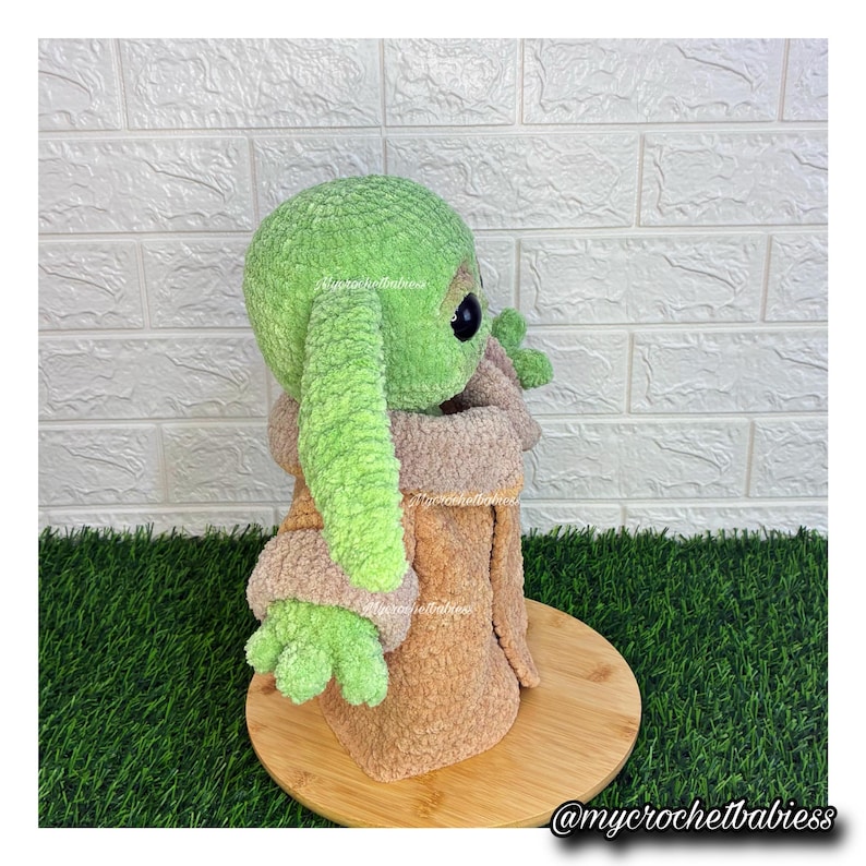 May include: A handmade green and tan plush doll of a character with large ears and big eyes. The doll is wearing a tan robe and is sitting on a wooden base. The doll is made of soft, textured yarn. The doll is a Star Wars inspired character.