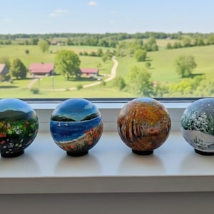 Op de afbeelding: Vier handgeschilderde decoratieve bollen met landschappen. Elke bol heeft een uniek ontwerp: een bloemenveld, een kustlandschap, een herfstbos en een besneeuwde winterscène. Ze worden tentoongesteld op een witte vensterbank.