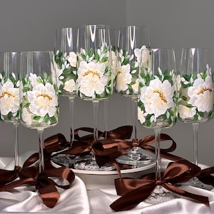 May include: A set of clear glass champagne flutes featuring hand-painted white peony flowers with green leaves and gold centres. Each stem is adorned with a brown satin ribbon tied in a bow. The base of the glasses features etched text.