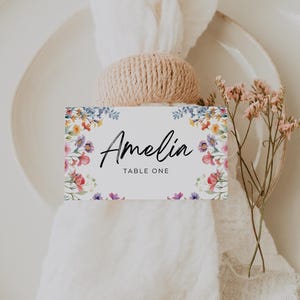 May include: A rectangular white place card featuring a colorful floral border with the name Amelia and the text TABLE ONE printed in black. The card is tucked into a tan woven napkin ring holding a white fabric napkin on a white plate.