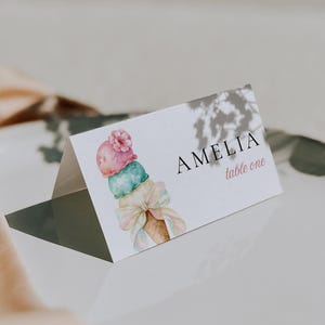 May include: A white folded place card featuring a watercolor illustration of an ice cream cone with a pink scoop, a teal scoop, and a light-colored bow. The text AMELIA is printed in black, with the words table one in pink script below it.