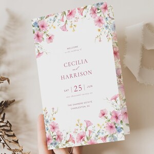 Könnte beinhalten: Eine Hochzeitseinladung mit floralen Akzenten. Die Einladung zeigt die Namen "Cecilia und Harrison" und das Datum "25. Juni". Das Design umfasst rosa und blaue Blumen. Ein weißes Band und getrockneter Farn sind ebenfalls vorhanden.