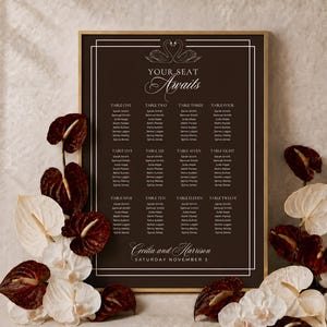 May include: A dark brown wedding seating chart sign in a gold frame featuring white text that reads Your Seat Awaits, a swan illustration, table numbers one through twelve with guest names, and the names Cecilia and Harrison with the date.