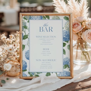 May include: A rectangular gold-toned frame containing a menu sign titled the BAR. The sign features blue hydrangea floral illustrations and lists wine, beer, and non-alcoholic selections in blue text on a white background with a blue border.