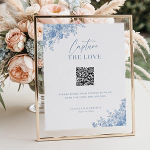 Op de afbeelding: Een ingelijst trouwbord met de tekst "Capture the Love" en een QR-code. Het bord heeft blauwe bloemenaccenten en de namen Cecilia & Harrison met de datum 15 juli 20XX. De lijst is goudkleurig.