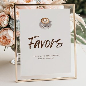 Op de afbeelding: Een ingelijst bord met het woord "Favors" in bruin script, bekroond met een illustratie van een koffiekopje. De tekst hieronder luidt "Neem iets om je dag op te fleuren!" De lijst is goudkleurig en de achtergrond is wit.
