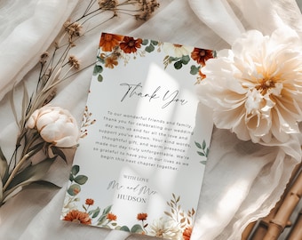 May include: A wedding thank you card with floral decorations in orange, red, and cream. The card has the words "Thank You" in elegant script and a personalized message. The card is surrounded by dried flowers and a large cream-colored flower.