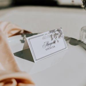 May include: A white place card with a black double-lined border and the name "Margaret Spencer" in elegant script. Two swan illustrations are at the top. The card is folded and sits on a white surface, with a peach-colored cloth in the background.