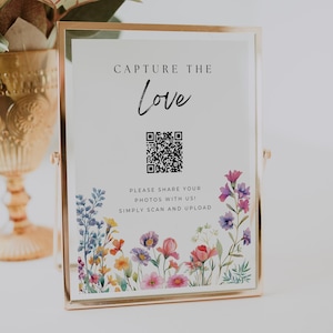 May include: A white sign with the text "Capture the Love" in black lettering. A black and white QR code is below the text. The text "Please share your photos with us! Simply scan and upload" is below the QR code. The sign is decorated with a watercolor floral border.