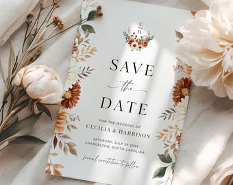 May include: A white 'Save the Date' card with floral accents. The card features the text 'SAVE the DATE' in elegant black font, along with wedding details. Floral elements in shades of brown, orange, and green frame the text.