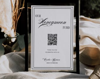 May include: A framed sign with the words "OUR Honeymoon FUND" in elegant script. Below is a QR code and the text "THANK YOU FOR CELEBRATING WITH US! Cecilia + Harrison 22ND OF OCTOBER 201X". The frame is black.