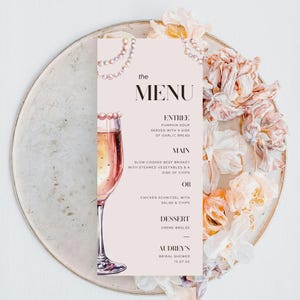 Könnte beinhalten: Eine rosa Speisekarte mit einem Glas Champagner und Perlenakzenten. Die Speisekarte enthält den Text "the MENU" und listet die Vorspeisen-, Hauptgericht- und Dessert-Optionen für eine Brautparty auf. Der Text am unteren Rand der Karte lautet "AUDREY'S BRIDAL SHOWER 15.07.XX".