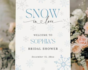 May include: A white sign on an easel with the words "SNOW in love" in blue script and snowflake designs. The sign welcomes guests to Sophia's bridal shower, with the date December 15, 20xx.