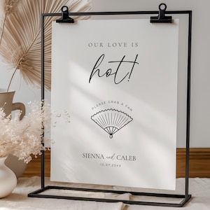 May include: A white sign with black text in a black metal frame. The sign reads "OUR LOVE IS hot! PLEASE GRAB A FAN" with a fan illustration. Names "Sienna and Caleb" and a date are at the bottom.
