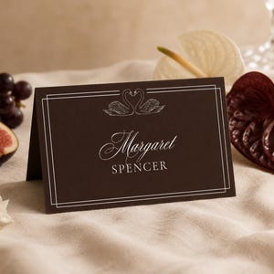 May include: A dark brown folded place card featuring a white line border, a graphic illustration of two swans forming a heart shape, and the printed text Margaret SPENCER in a combination of elegant script and clean sans-serif typography.
