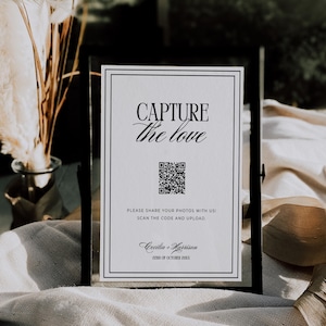 May include: A framed sign with the text "CAPTURE the love" in elegant script. Below the text is a QR code and the words "PLEASE SHARE YOUR PHOTOS WITH US! SCAN THE CODE AND UPLOAD." The sign also includes the names "Cecilia + Harrison" and the date "22ND OF OCTOBER 20XX".