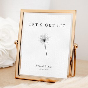 May include: A gold-framed sign with the text "LET'S GET LIT" above a black and white dandelion illustration. Below the illustration are the names "AVA and LIAM" and the date "July 15, 20xx". The sign is displayed on a wooden surface.