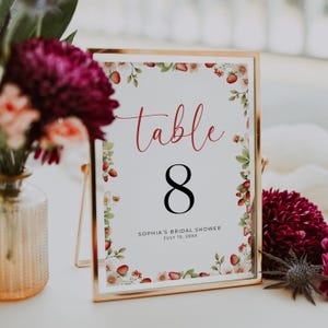 May include: A gold framed table number 8 sign with a floral strawberry design. The word "table" is in red script above the number 8. The text "Sophia's Bridal Shower July 15, 20xx" is below the number.