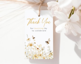 May include: A rectangular white paper gift tag hanging from a tan twine string. The tag features gold cursive text reading Thank You, black text reading for buzzing by to celebrate, and a watercolor illustration of yellow wildflowers and bees.
