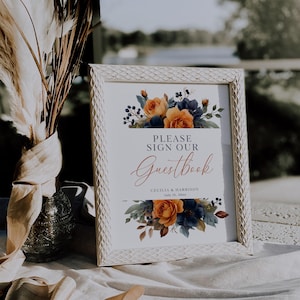 May include: A framed wedding guestbook sign with the text "PLEASE SIGN OUR Guestbook" in elegant script. The sign features floral arrangements in orange, blue, and green. The frame is white with a textured pattern. The names "Cecilia & Harrison" and the date are also included.