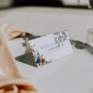 Op de afbeelding: Een witte buffetkaart met blauwe en oranje bloemmotieven. De kaart toont de tekst "BUFFET CARDS" en "editable text", met de slogan "CREATE UNLIMITED CARDS". De kaart is gevouwen en ligt op een wit oppervlak.
