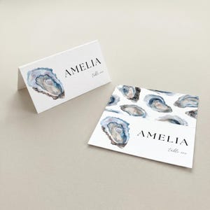 May include: White folded place cards with watercolor oyster shell illustrations. The name "AMELIA" is printed in black, with "table one" below. One card is folded, the other is flat with multiple oyster shell designs.