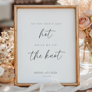 May include: A framed sign with gold trim and white background. The sign reads "So you don't get hot while we tie the knot" in elegant black script. Names and a date are at the bottom. Floral arrangements are in the background.